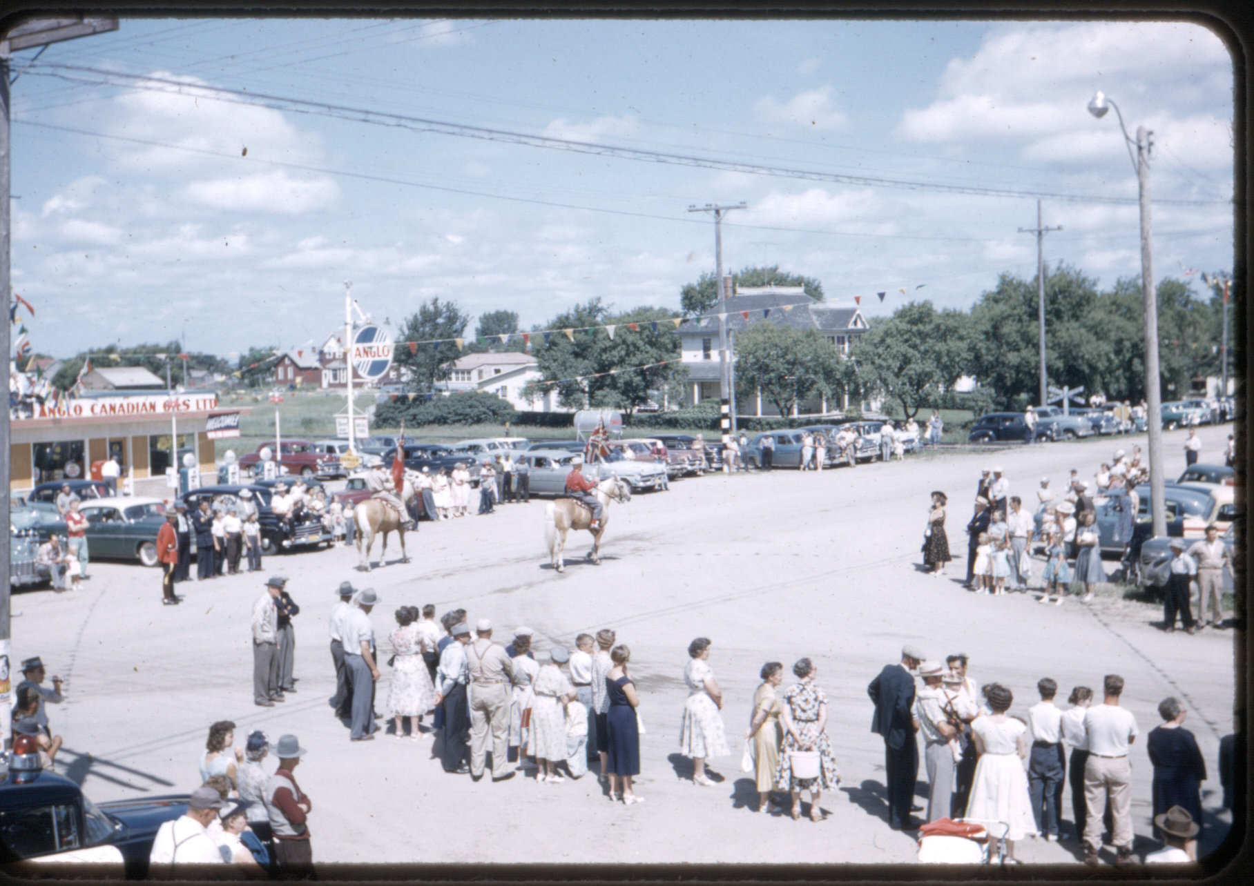 [Scanned photo of the Mill Road/South Railway intersection during a parade showing the Anglo-Canadian Oils fuel station on the west side next to the railway]
